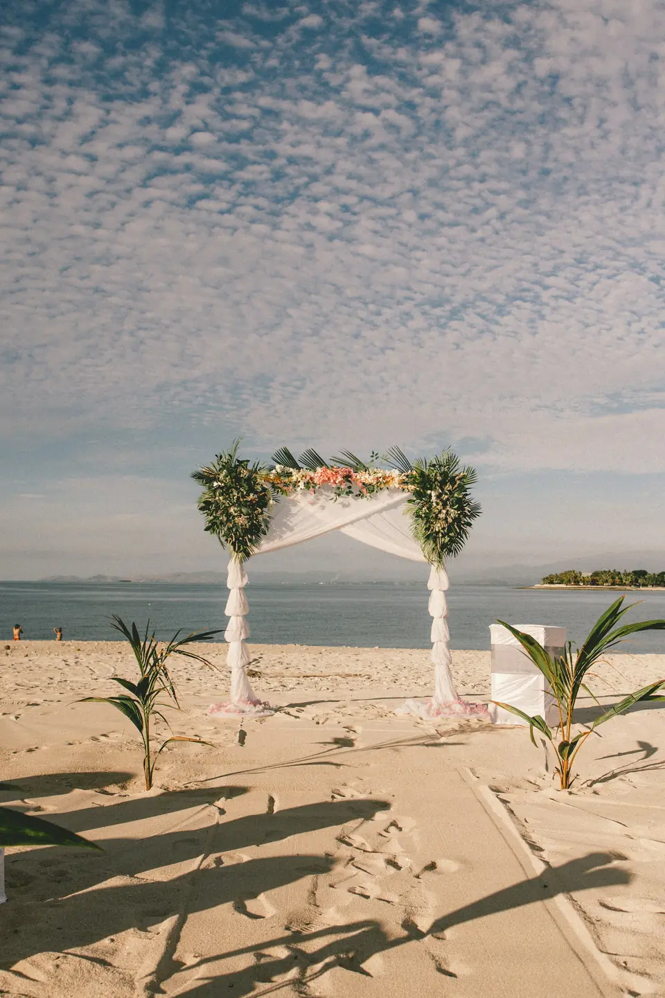 A floral ceremony arch standing on the sand with palm leaves