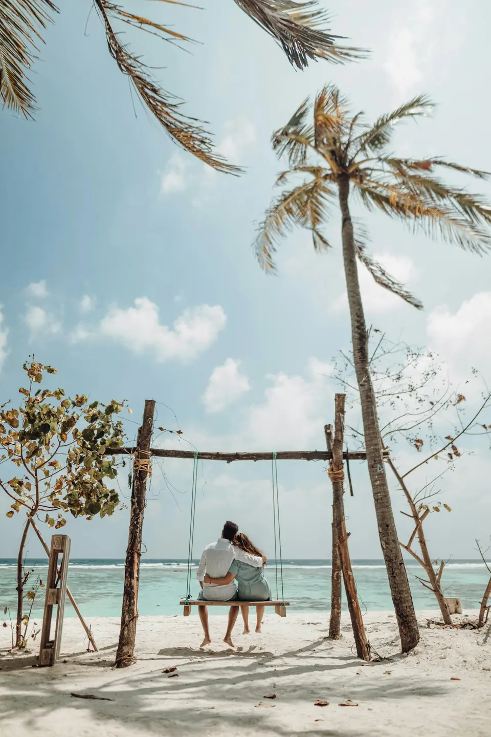 A couple on a swing beneath palm trees on a white beach