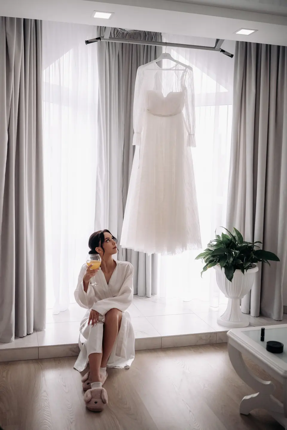 A bride in a robe sitting beneath her hanging dress while getting ready