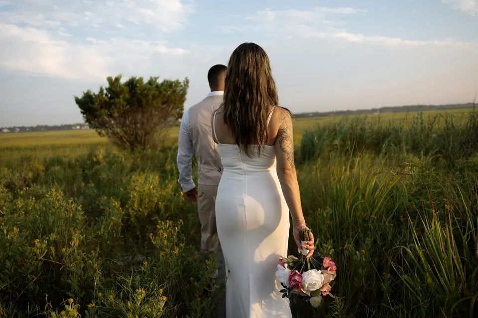 A bride and groom walking through a grassy field in the evening light