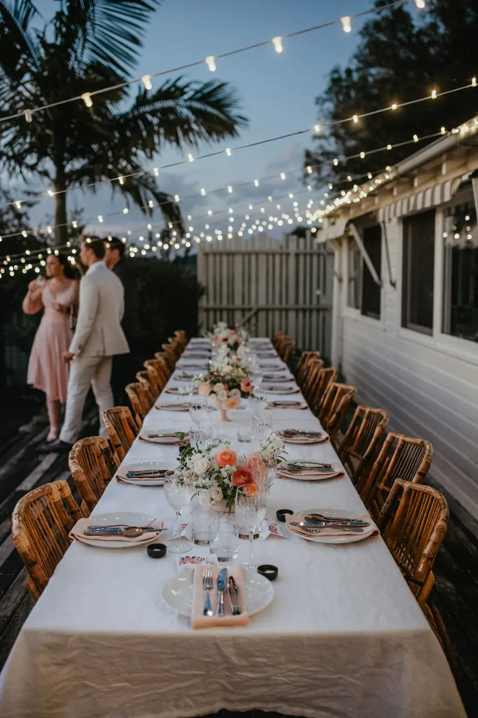 A couple standing at the head of a long table under string lights and palms