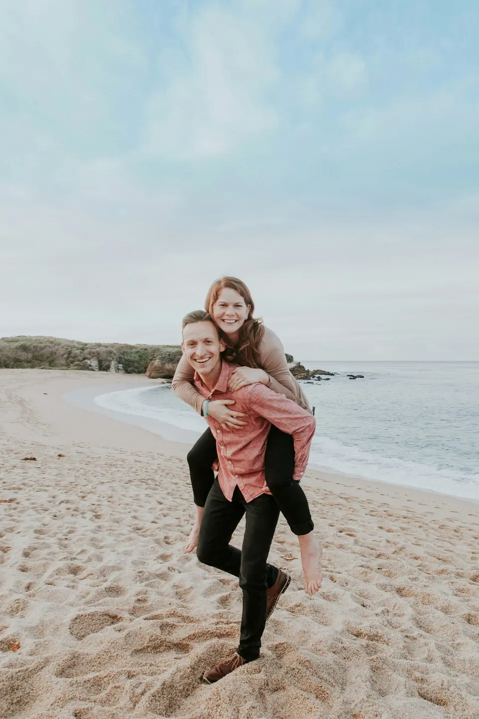 A laughing couple doing a piggyback on the beach for engagement photos
