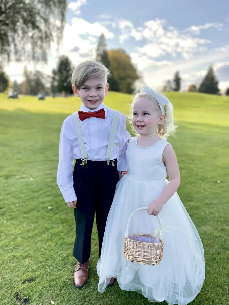 A ring bearer in a red bow tie and a flower girl with a basket on a lawn