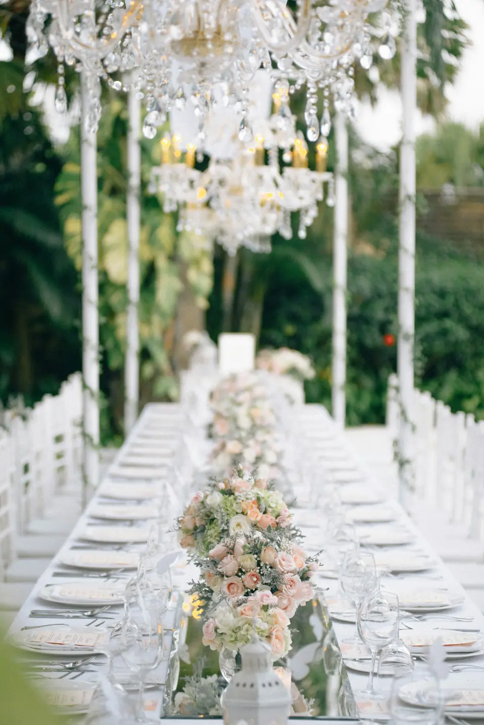 A long reception table with a flower runner under crystal chandeliers outdoors