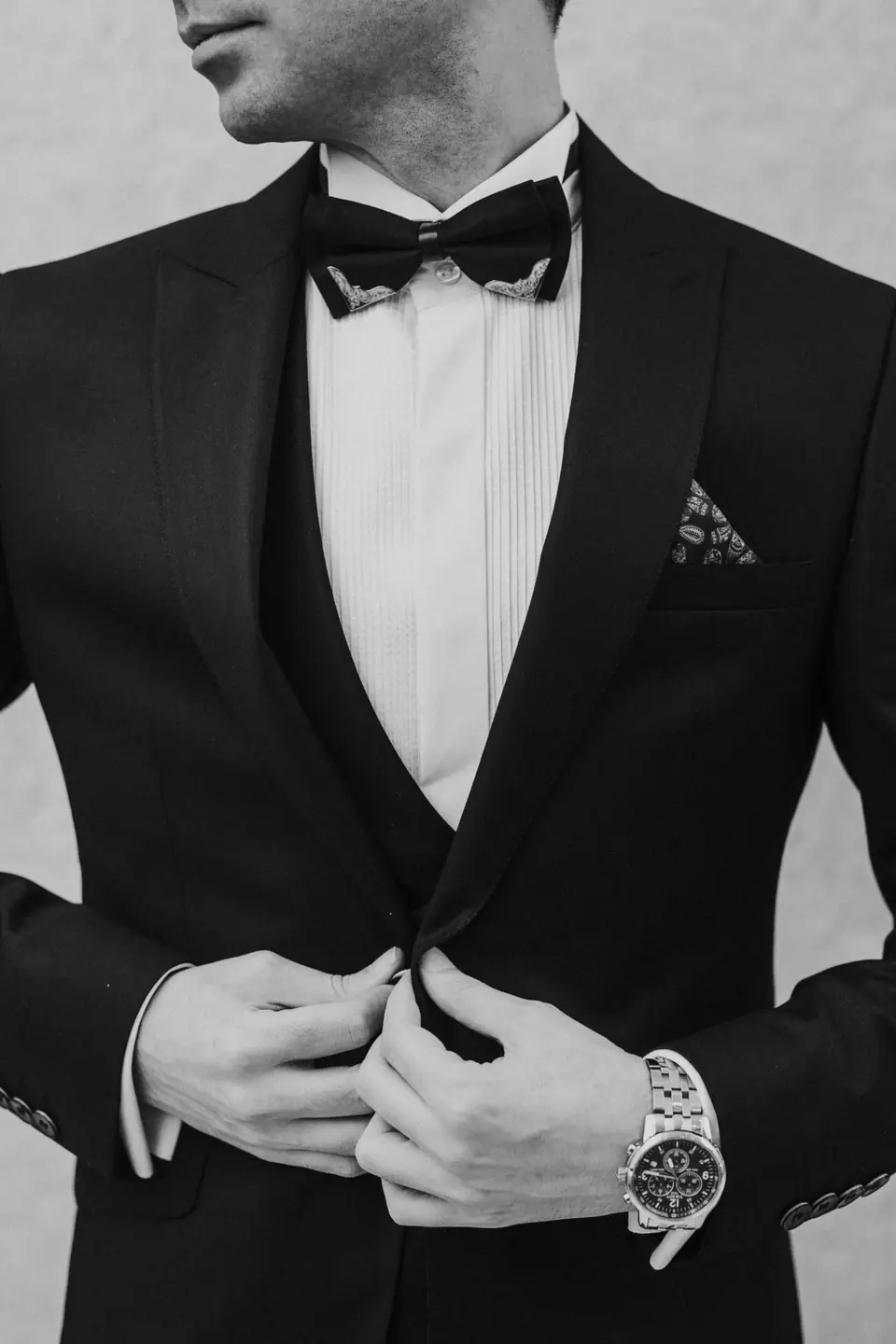 A groom buttoning a black tuxedo jacket, in black and white